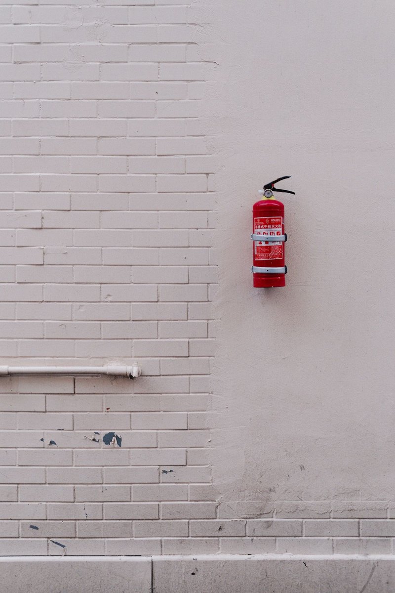 Red fire extinguisher mounted on a pale painted brick wall beside a white pipe