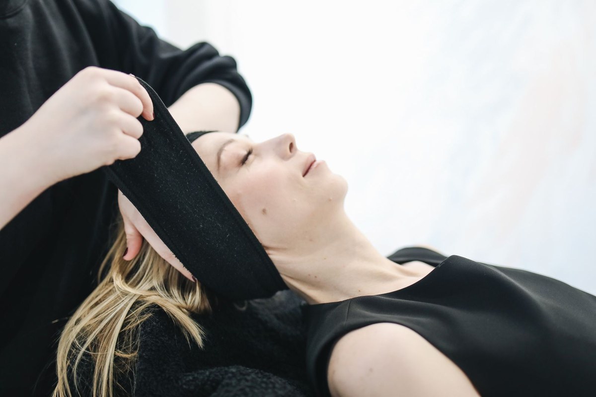 Woman with closed eyes on a treatment bed while a therapist settles a black headband across her forehead